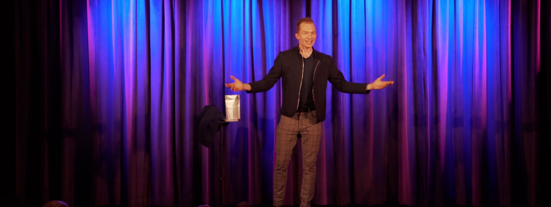 Trigg Watson on stage with arms wide open in front of blue and purple curtains