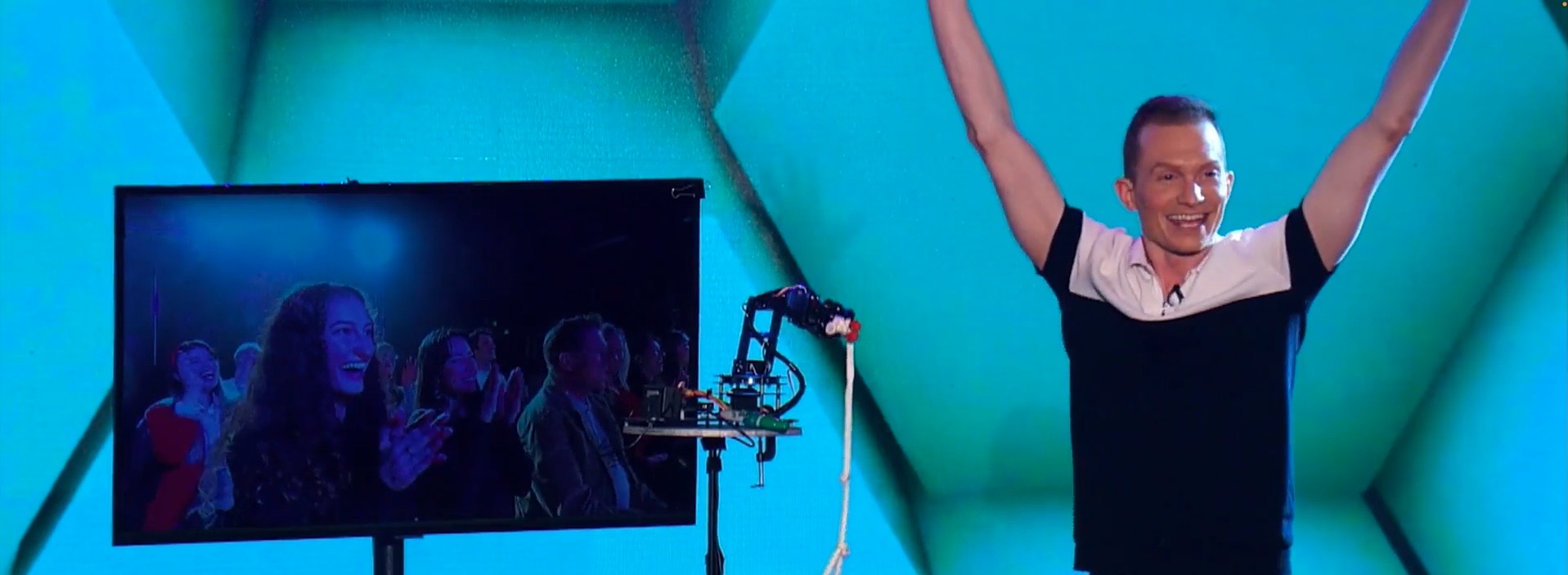 Trigg Watson celebrating on stage with a robot arm and a screen showing the amazed audience